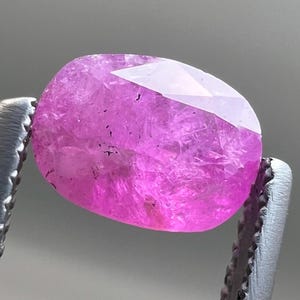 May include: A faceted, oval-shaped pink gemstone held by metal tweezers. The gem has a vibrant, saturated pink color with internal inclusions and a polished surface. The tweezers are silver-colored, and the background is a neutral gray.