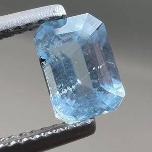 Puede incluir: Una gema de aguamarina rectangular facetada, sujeta por unas pinzas metálicas. La gema es de color azul claro, con estructuras internas y facetas visibles. Las pinzas son de color gris oscuro.