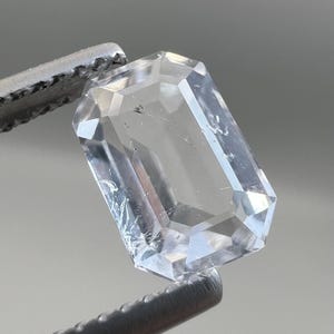 Può includere: Una gemma chiara e rettangolare con molteplici sfaccettature, tenuta da uno strumento di metallo. La gemma ha un aspetto trasparente, riflettendo la luce e mostrando il suo taglio. La gemma è di un colore chiaro e neutro.