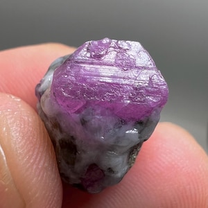 May include: A close-up of a rough, uncut ruby gemstone. The ruby is a vibrant, deep pink color with a crystalline structure. The stone is set against a neutral background, highlighting its natural beauty.