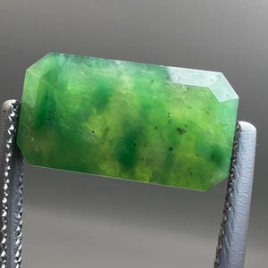 Pode incluir: Uma pedra preciosa retangular e facetada com um tom verde vibrante, segurada por uma ferramenta de metal prateado. A cor da pedra preciosa varia de verde claro a escuro, com manchas mais escuras. As bordas são cortadas com múltiplas facetas.
