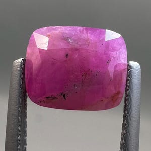 May include: A faceted, cushion-cut ruby gemstone. The gem is a vibrant pinkish-red color, held by silver tweezers. The stone has a polished surface with visible internal inclusions and a high degree of transparency, reflecting light.
