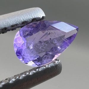 May include: A faceted, pear-shaped gemstone in shades of lavender and purple is held by a pair of silver tweezers. The gem's surface reflects light, showcasing its cut and clarity. The background is a neutral gray.
