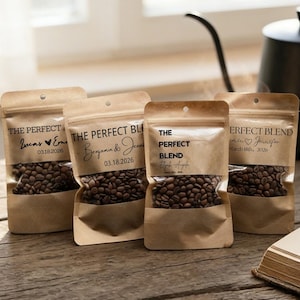May include: Four personalized coffee bean bags with the text "THE PERFECT BLEND" and names and dates. The bags are brown with a clear window showing the coffee beans. A coffee pot, a cup of coffee, and a book are in the background.