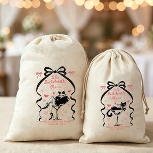 May include: Two off-white drawstring canvas bags featuring a black and pink graphic print. The design includes a bow, a martini glass, and text reading The Bachelorette Eliana and Eliana's Bachelorette Party London 10.15.25 with small hearts.