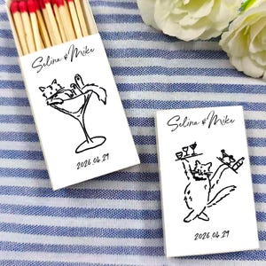 May include: White matchbox with red-tipped matches. The box features a black line drawing of a cat in a martini glass and the text "Selina & Mike" and "2026.04.29". Another box shows a cat holding a tray.