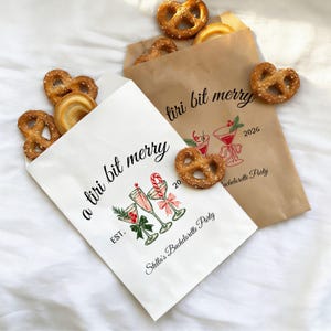 Op de afbeelding: Twee papieren zakjes, een witte en een bruine, gevuld met zoute krakelingen. Het witte zakje bevat de tekst 'a tiri bit merry', 'EST.' en 'Stella's Bachelorette Party' met illustraties van drie cocktailglazen en een zuurstok.