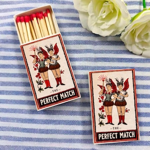 May include: Open matchbox with red-tipped matches and a closed matchbox. Both boxes feature a vintage illustration of two winged figures with the text "~THE~ PERFECT MATCH" in a red border. The background is a blue and white striped fabric.