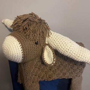 May include: A handmade crochet Highland cow toy in shades of brown and cream. The toy features long, cream-colored horns, a shaggy mane, and a detailed body. The cow is designed with a textured stitch pattern and black button eyes.