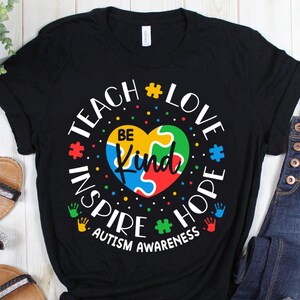 May include: Black t-shirt with a white graphic design that says "Teach Love Inspire Hope Be Kind" with a jigsaw piece heart in the centre. The text is surrounded by jigsaw pieces and the words "Autism Awareness" are at the bottom.