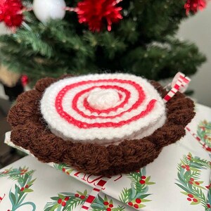 Op de afbeelding: Een gehaakt kerstboom ornament in de vorm van een yule log met een bruine chocolade basis en een wit en rood swirl ontwerp. Het ornament ligt op een ingepakt cadeau met een feestelijk ontwerp.