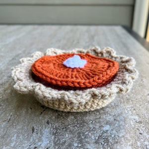May include: A crocheted pumpkin pie with a white dollop of whipped cream on top. The pie is brown and orange and has a ruffled crust.