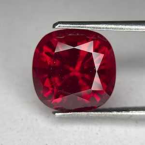 May include: A cushion-cut, deep red gemstone held by tweezers. The gem has a faceted surface that reflects light, showcasing its brilliance. The color is a rich, saturated red, typical of a ruby. The gem is likely used in jewelry.