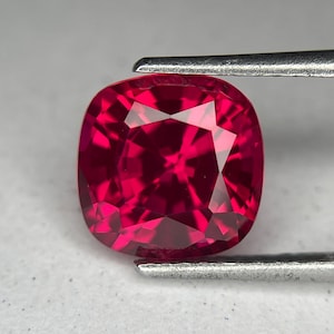 May include: A vibrant, cushion-cut gemstone, held by tweezers. The gem is a deep, saturated red, with multiple facets reflecting light. The shape is a rounded square, showcasing the stone's brilliance.