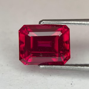 May include: A faceted, rectangular-cut ruby gemstone held by silver tweezers. The ruby is a deep, saturated red color with internal reflections. The gemstone's facets create a brilliant display of light and shadow.