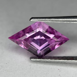 May include: A faceted, kite-shaped gemstone in shades of pink and purple is held by metal tweezers. The gem has a symmetrical, geometric cut, reflecting light and showcasing its internal structure. The gem's color ranges from light to deep pink.
