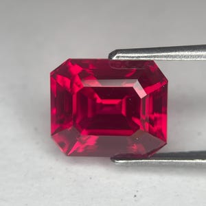 Könnte beinhalten: Ein facettierter, rechteckig geschliffener Rubin-Edelstein. Der Edelstein hat eine tiefe, satte rote Farbe und wird von einer Pinzette gehalten. Der Schliff des Steins zeigt mehrere Facetten, die seine Brillanz und Klarheit verbessern. Der Edelstein ist ein Blickfang und zeigt seine satte Farbe und Form.