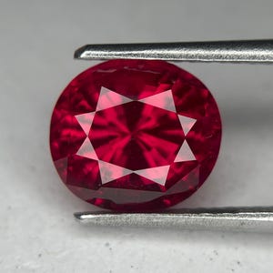 Può includere: Una gemma rotonda, sfaccettata, di colore rosso intenso, tenuta da pinzette metalliche. La gemma presenta un motivo a stella di sfaccettature che riflettono la luce. Il colore è un rosso vibrante e saturo, con una superficie lucida.