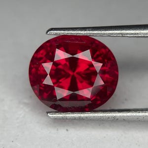 May include: A faceted, round-cut, deep red gemstone held by metal tweezers. The gem has a star-like pattern of light reflections. The gemstone is a vibrant, saturated red color.