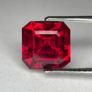 May include: A faceted, square-cut, deep red gemstone. The gem is held by a pair of silver tweezers, showcasing its brilliant facets and internal reflections. The gemstone's color is a rich, saturated red.