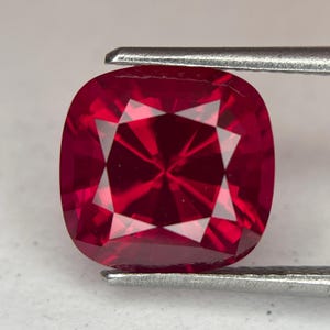 May include: A cushion-cut, faceted ruby gemstone held by a pair of silver tweezers. The ruby is a deep, saturated red color with a brilliant, star-like pattern in the center. The gemstone's facets reflect light, creating a sparkling effect.