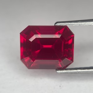May include: A faceted, rectangular ruby gemstone held by tweezers. The gem is a deep, saturated red color with sharp, clean edges and a polished surface. The cut of the stone allows light to reflect, creating a brilliant appearance.