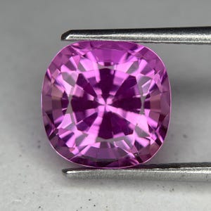 Könnte beinhalten: Ein facettierter, kissenförmiger, rosa Edelstein, der von einer Metallpinzette gehalten wird. Das Innere des Edelsteins zeigt ein Sternmuster aus hellen und dunklen rosa Facetten. Der Edelstein hat einen leuchtenden Rosaton.