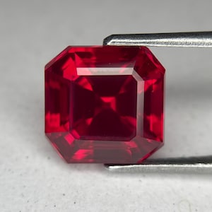 May include: A radiant, square-cut ruby gemstone, held by silver tweezers. The gem exhibits a deep red colour with facets that reflect light. The gemstone's shape is an octagonal square, highlighting its clarity and brilliance.