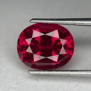 May include: A faceted, oval-shaped ruby gemstone held by tweezers. The ruby is a deep, rich red color with a starburst pattern of facets reflecting light. The gemstone is held against a neutral background.