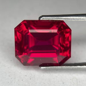 May include: A faceted, rectangular-cut ruby gemstone held by a silver metal tool. The ruby is a deep, vibrant red color with a reflective surface. The gemstone's edges are precisely cut, showcasing its internal structure.
