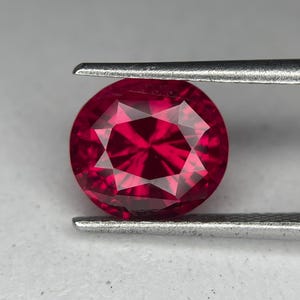May include: A round, faceted ruby gemstone held by a pair of tweezers. The gemstone is a deep, vibrant red color with a starburst pattern visible in the center. The tweezers are silver.