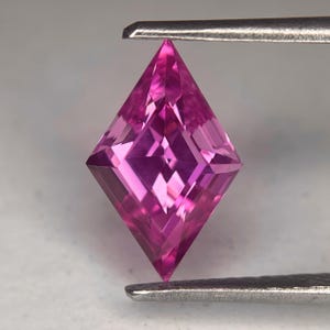 May include: A faceted, pink gemstone held by metal tweezers. The gem is cut in a kite shape, with multiple facets reflecting light. The color is a vibrant, medium pink, and the gem appears to be transparent.