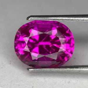 Könnte beinhalten: Ein facettierter, oval geformter, pinkfarbener Edelstein, der von einer silbernen Pinzette gehalten wird. Der Edelstein hat mehrere Facetten, die das Licht reflektieren und eine brillante Farbdarstellung erzeugen. Der Edelstein ist ein leuchtendes Pink.