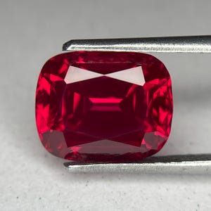 May include: A cushion-cut, faceted ruby gemstone held by tweezers. The ruby is a deep, saturated red color with multiple facets that reflect light. The gemstone is a square shape with rounded corners.
