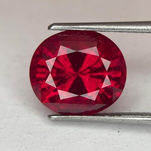 May include: A faceted, round-cut ruby gemstone held by tweezers. The gem is a deep, vibrant red with internal facets creating a starburst pattern. The tweezers are silver-colored, and the background is a neutral white.