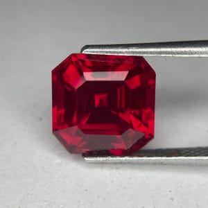 May include: A faceted, square-cut, deep red gemstone held by tweezers. The gem has a symmetrical, multi-faceted design, reflecting light. The color is a rich, saturated red, with a square-shaped center. The gem is likely a ruby.