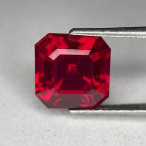 Può includere: Un rubino sfaccettato a taglio quadrato, tenuto da una pinzetta. La gemma è di un colore rosso intenso e saturo, con sfaccettature nitide e pulite che riflettono la luce. Il rubino è di forma quadrata.