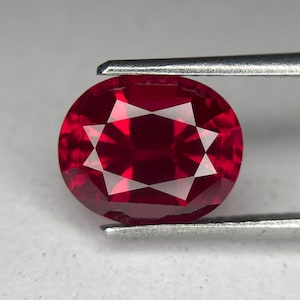 May include: A faceted, oval-cut ruby gemstone held by tweezers. The gem is a deep, vibrant red with internal light reflections. The facets create a star-like pattern in the center. The tweezers are silver-colored.