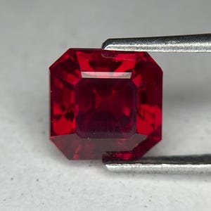 May include: A faceted, deep red gemstone, held by a pair of silver tweezers. The gem has an octagonal shape with multiple facets that reflect light. The stone's rich color and cut suggest it could be a ruby.