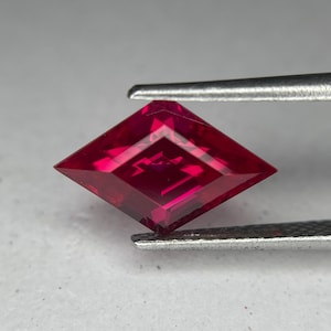 May include: A faceted, kite-shaped ruby gemstone held by tweezers. The gem is a deep, saturated red with internal facets that reflect light. The tweezers are silver-colored, and the background is a neutral gray.