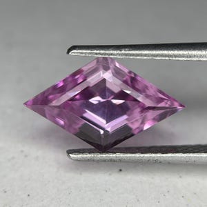 May include: A faceted, kite-shaped gemstone in shades of pink and purple is held by a pair of tweezers. The gem has a symmetrical cut with multiple facets that reflect light, creating a sparkling effect. The gemstone's color ranges from light to deep pink.