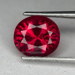 Può includere: Una gemma rotonda, sfaccettata, rosso intenso, tenuta da pinzette d'argento. La gemma presenta un motivo a stella di luce che si riflette all'interno della sua superficie lucida. La gemma è di un colore rosso vibrante e saturo.