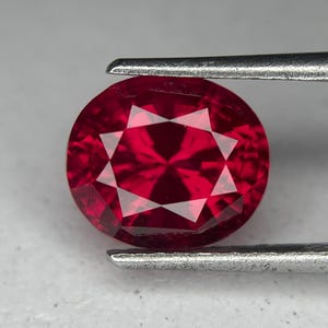 May include: A round, faceted ruby gemstone held by tweezers. The gem is a deep red colour with internal facets creating a starburst pattern. The tweezers are silver, and the background is a neutral grey.