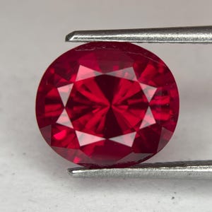 May include: A round, faceted ruby gemstone held by metal tweezers. The ruby is a deep, vibrant red color with multiple facets that reflect light. The gemstone is round and appears to be polished, with a brilliant cut.