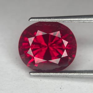 May include: A faceted, round-cut ruby gemstone held by tweezers. The gemstone is a deep, vibrant red color with multiple facets that reflect light. The tweezers are silver-colored and hold the gemstone against a white background.
