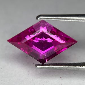 May include: A faceted, magenta-colored gemstone held by tweezers. The gemstone is cut in a kite shape, with multiple facets reflecting light. The color is a deep, vibrant pink, with a slight variation in tone across the surface.