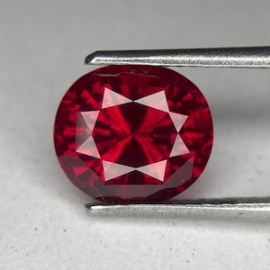 May include: A round, faceted, deep red gemstone held by metal tweezers. The gem has a star-like pattern in the center, reflecting light. The stone's color is a vibrant, saturated red, and it appears to be a ruby.