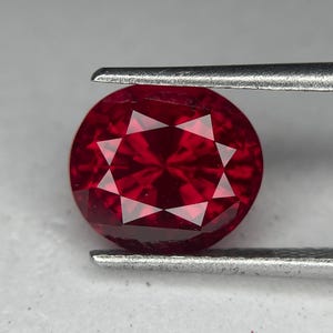 Può includere: Una gemma rotonda, sfaccettata, rosso intenso, tenuta da una pinzetta d'argento. La gemma ha un motivo a stella di sfaccettature che riflettono la luce. La gemma è di un colore vibrante e saturo, e le sfaccettature creano un effetto scintillante.