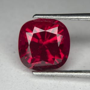 May include: A cushion-cut, faceted ruby gemstone. The gem is a deep, saturated red color, held by a pair of silver tweezers. The facets create a brilliant display of light and shadow, highlighting the gem's clarity and shape.
