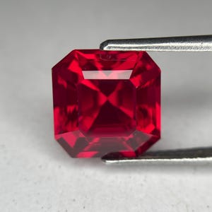 May include: A faceted, square-cut ruby gemstone held by tweezers. The gem is a deep, vibrant red with sharp, reflective facets. The gemstone's shape is a modified square with cut corners, showcasing its brilliance.